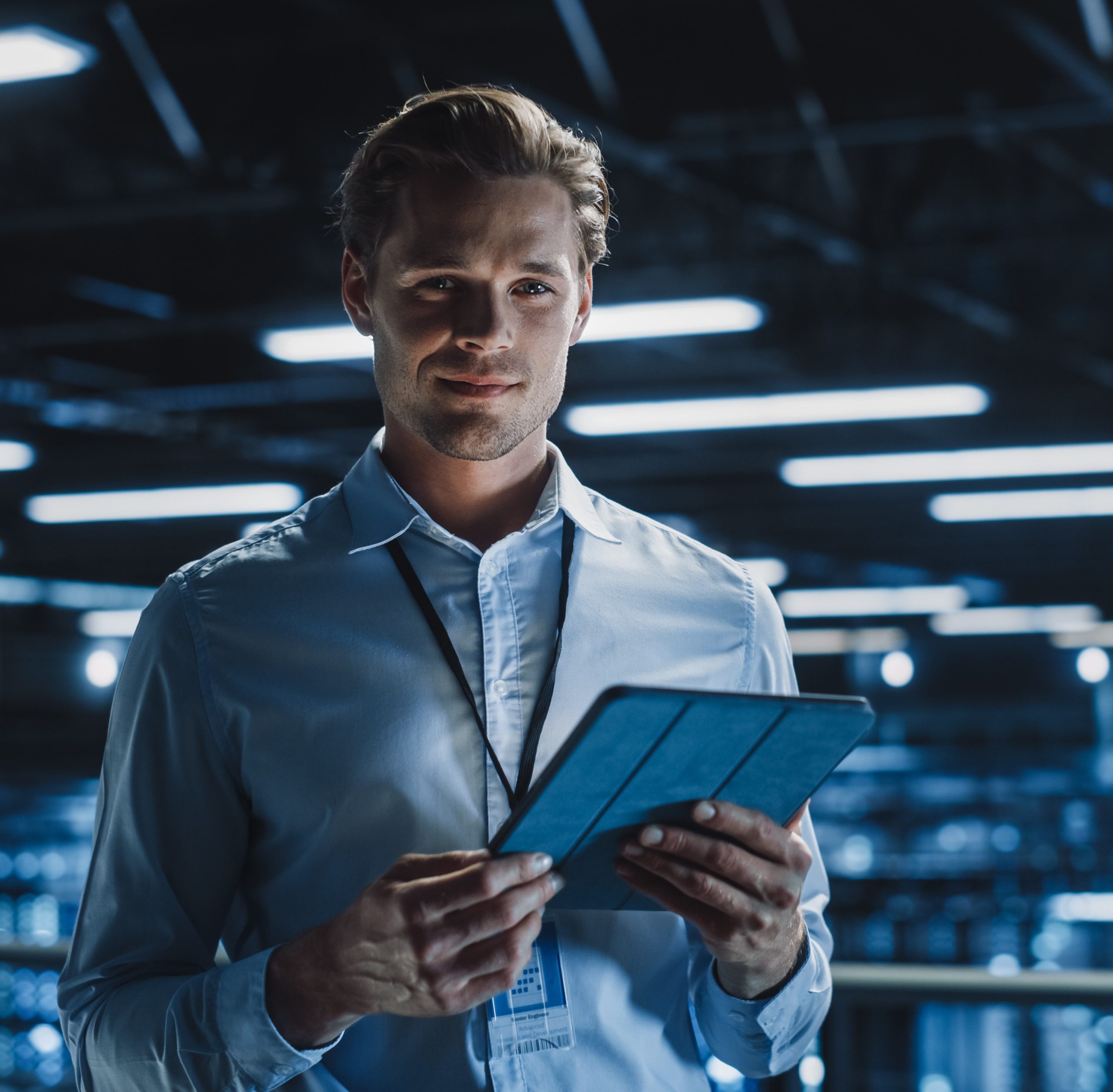 Portrait of Handsome Smiling IT Specialist Using Tablet Computer in Data Center, Looking at Camera. Succesful Male e-Business Engineer Working in Big Server Farm Cloud Computing Facility.
